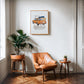Oak-framed orange BMW 2002 artwork hanging above a tan leather chair and potted plants.