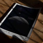 Unframed Artemis II art print showing a crescent Earth above the cratered lunar horizon, lying on a wooden table in dramatic shadow.