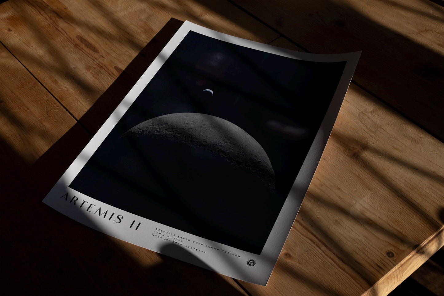 Unframed Artemis II art print showing a crescent Earth above the cratered lunar horizon, lying on a wooden table in dramatic shadow.