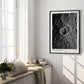 Black-framed Artemis II lunar crater art print hanging in a sunlit room above a window bench with cushions, books, and flowers.