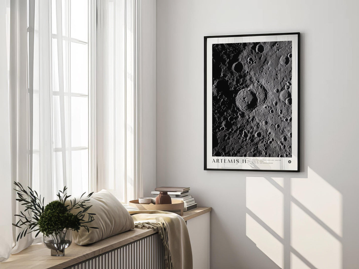 Black-framed Artemis II lunar crater art print hanging in a sunlit room above a window bench with cushions, books, and flowers.