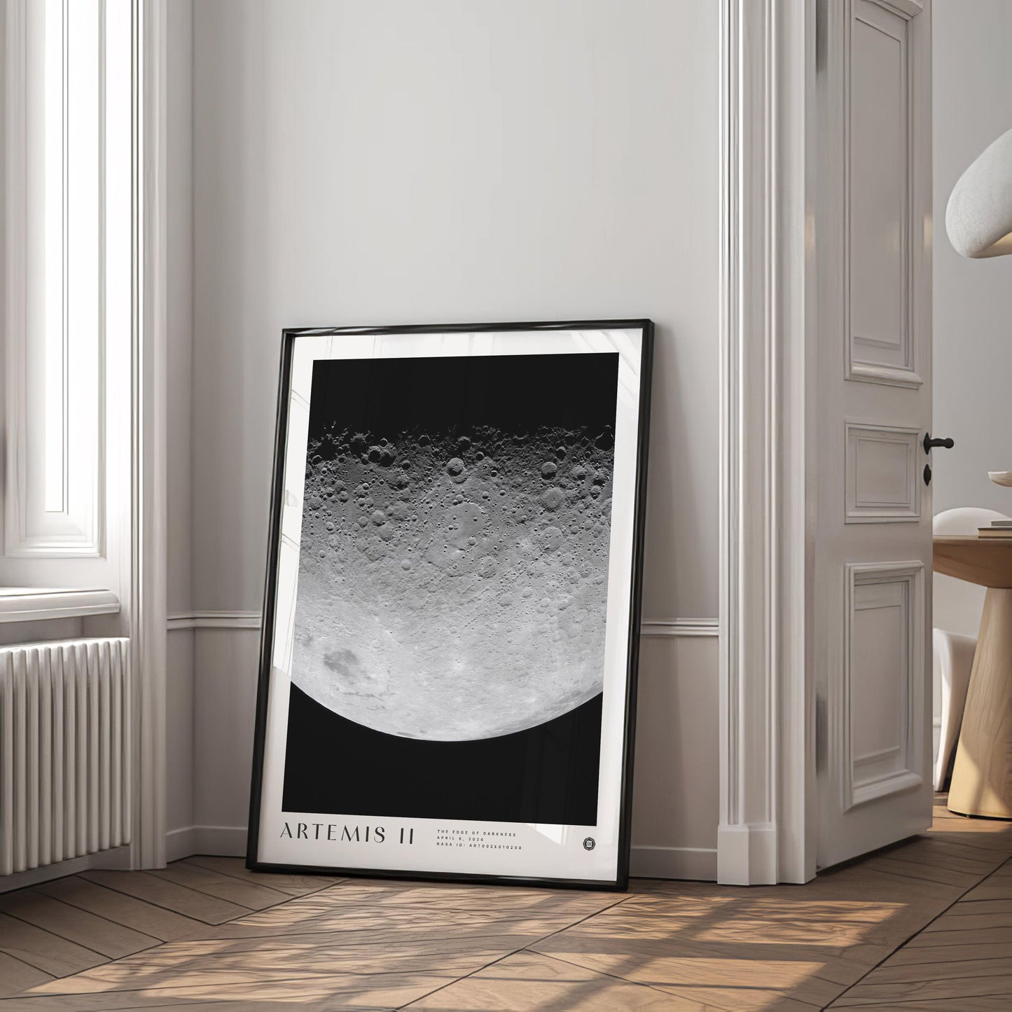 Black-framed Artemis II lunar surface art print leaning beside a window in a bright interior.