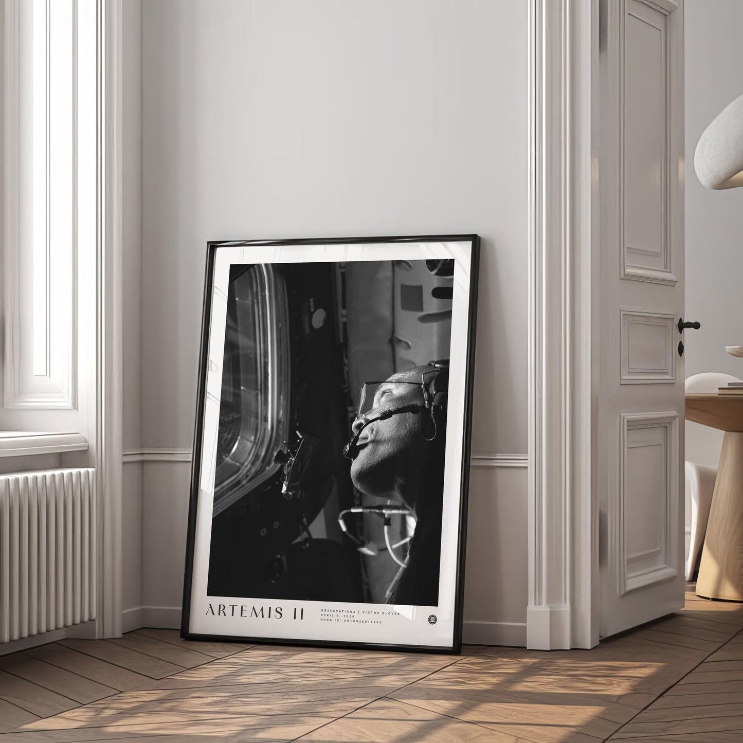 Black-framed Artemis II astronaut art print leaning against the wall in a bright room beside a window and open doorway.