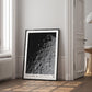 Black-framed Artemis II lunar surface art print leaning against a wall in a bright room beside a window and open doorway.