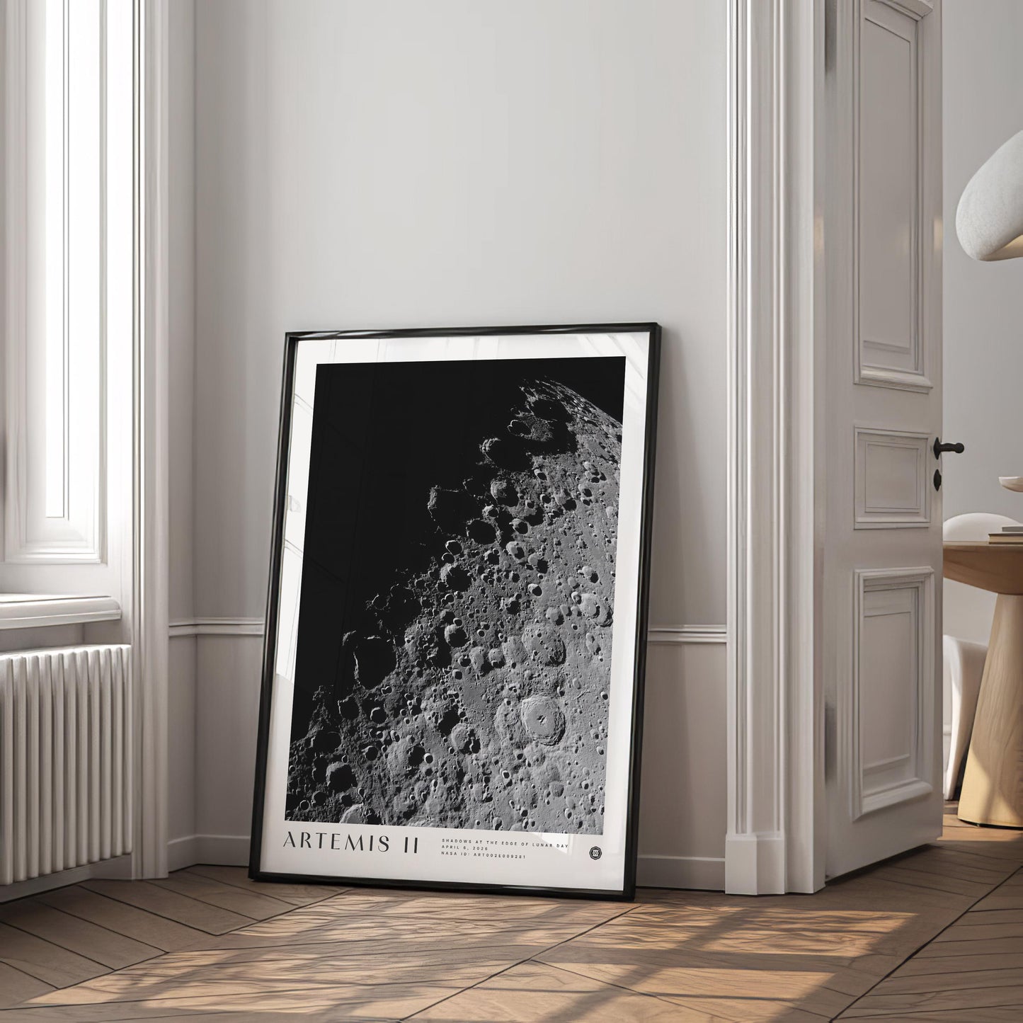 Black-framed Artemis II lunar surface art print leaning against a wall in a bright room beside a window and open doorway.