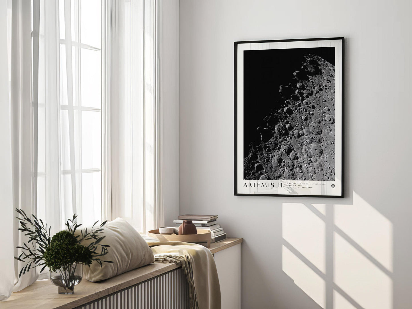 Black-framed Artemis II lunar surface art print hanging in a sunlit room above a window bench with cushions, books, and flowers.
