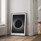 Black-framed Artemis II eclipse artwork leaning on a wooden floor beside a bright window and doorway.