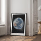 Black-framed Artemis II Earth artwork leaning on a wooden floor beside a bright window and doorway.