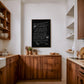 Dark wood-framed black dining-themed artwork hanging above wooden kitchen cabinets.