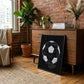 Black-framed soccer diagram artwork leaning against a wooden dresser in a room with exposed brick, plants, and wood flooring.