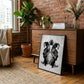 Black-framed monochrome DJ turntable artwork leaning against a wooden cabinet in a brick-walled room with plants and books.